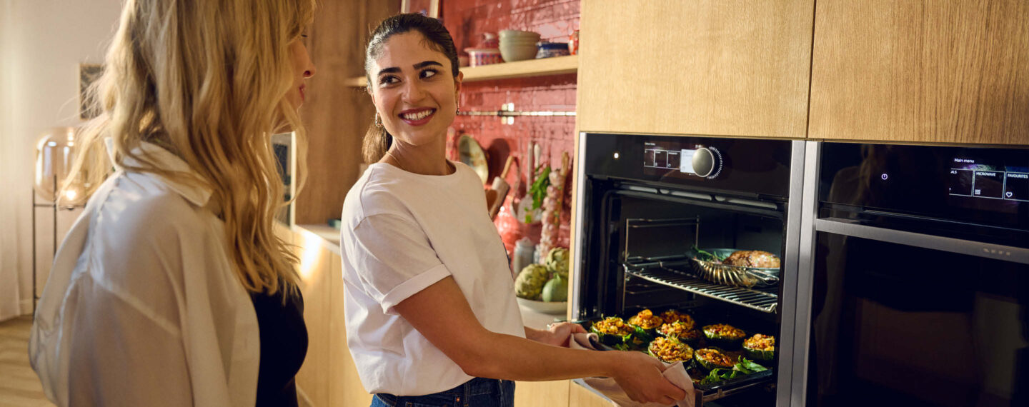 „Moderne Einbauküche mit hochwertigen NEFF Geräten, zwei Frauen beim gemeinsamen Kochen, geöffneter Backofen mit frisch zubereiteten Speisen und warmem Lichtambiente