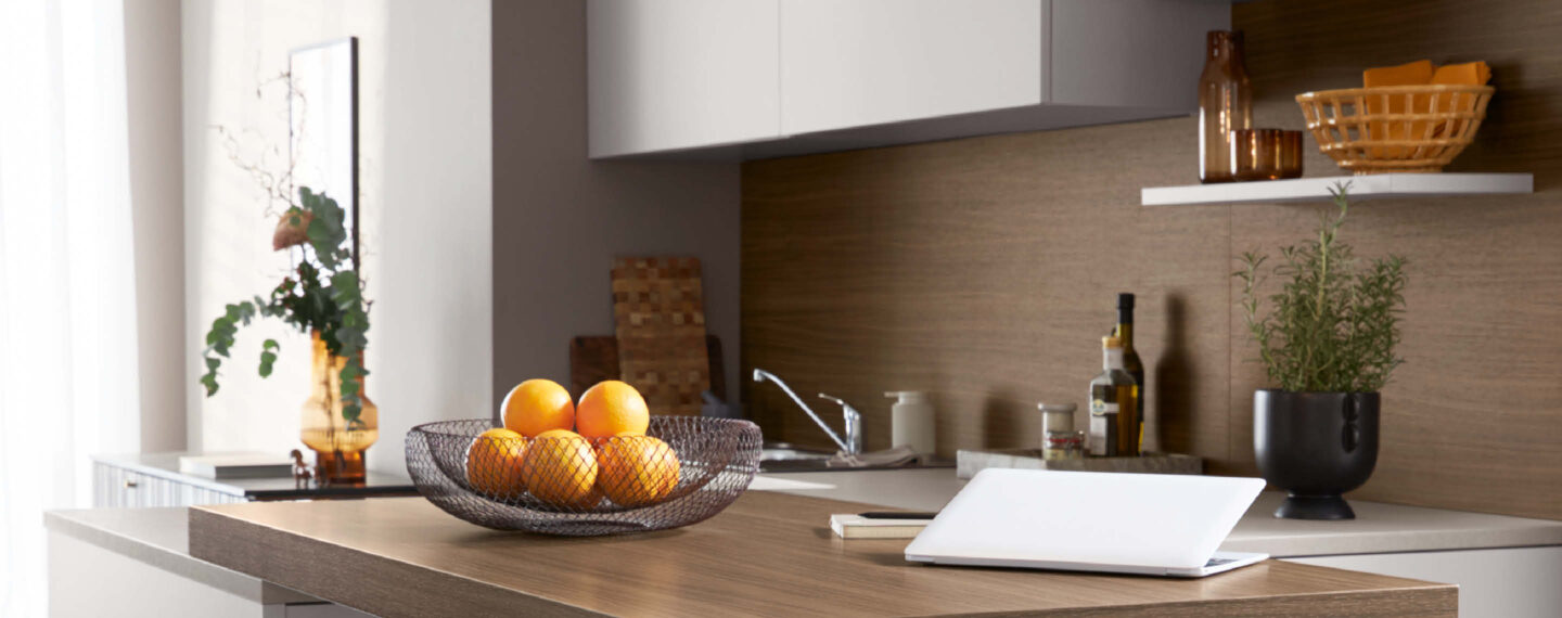 Moderne Schüller Küche mit heller Arbeitsplatte, Obstschale mit Orangen und stilvollen Accessoires, kombiniert mit natürlicher Holzrückwand und wohnlicher Atmosphäre